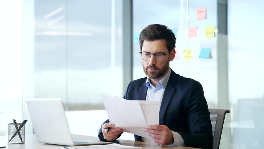 Busy serious financier working checks documents using a laptop computer in modern business office. Thoughtful businessman in formal suit is doing paperwork while sitting at the desk at the workplace