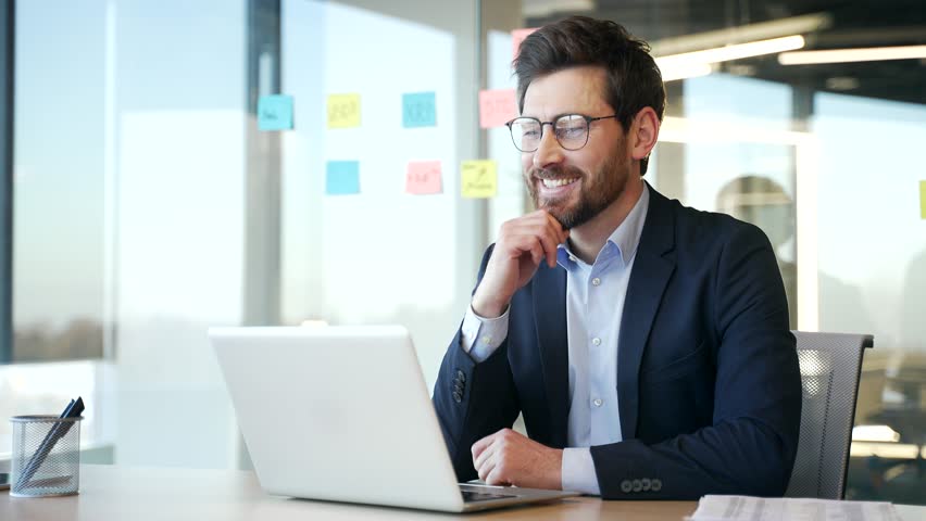Businessman working at desk in modern office using laptop. Smiling man focused, planning business strategy in contemporary workplace. Manager in glasses working in computer application, banking