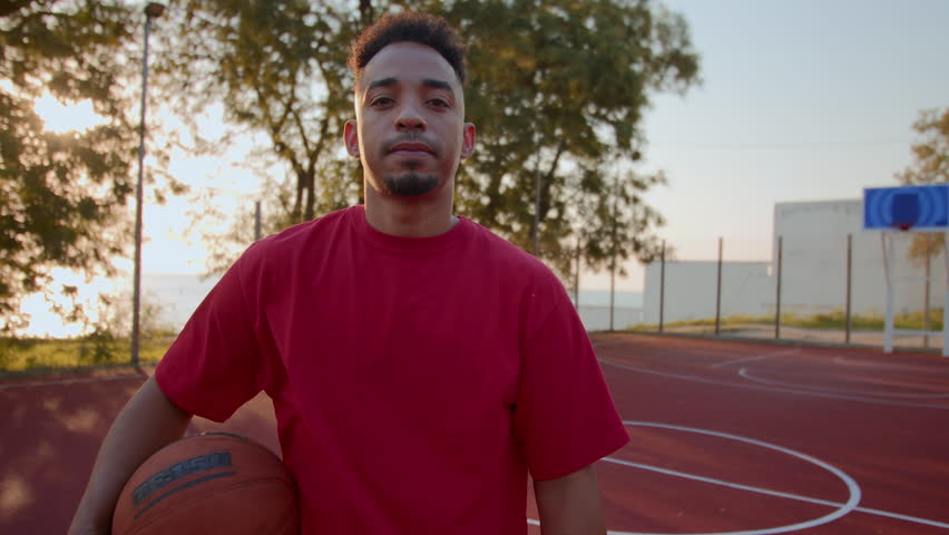 Confident young man holds ball standing at outdoor basketball court on summer day. Black sportsman in red t-shirt reflects readiness to play active game