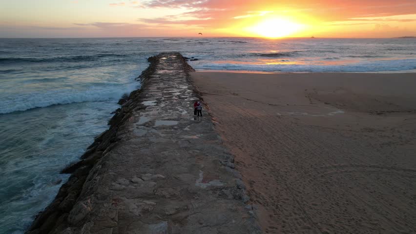 A married couple look at the waves crashing on the pier. Male and female contemplating the spray of waves crashing on a rocky shore during sunset. Couple standing and looking out over Atlantic Ocean.