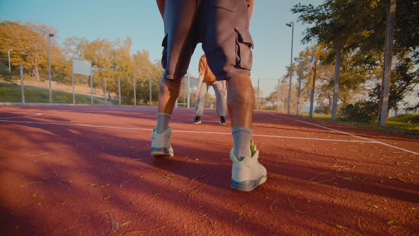 Sportive young black man dribbles the ball running along outdoor court with teammates on sunny day. Active people engage in playing basketball game together