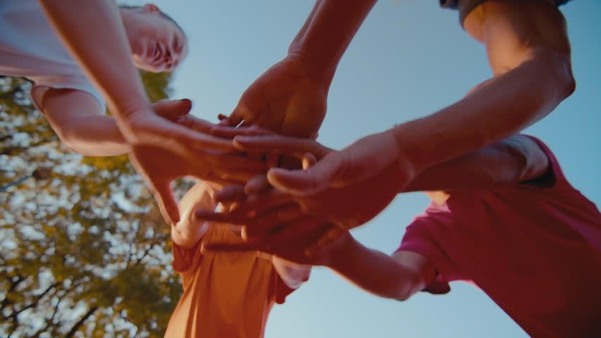 Cheerful people gather putting hands together against sky. Women and men in sportswear engage in team building activity before playing basketball