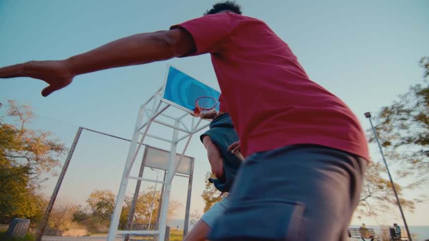 Young sportsmen fight for ball playing basketball on playground in evening. African American man makes jump shooting ball into hoop without efforts