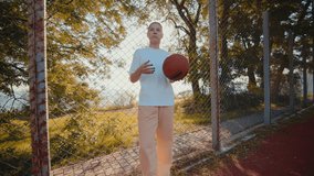 Smiling young woman holds ball putting from hand to hand against fence on playground. Cheerful lady basketball player reflects confidence with hobby at back sunset - Powered by Shutterstock - Get 15% off with code: PIKWIZARD15