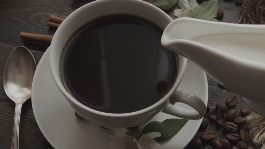 Pouring cream into black coffee in cup, with cinnamon sticks, coffee beans and flowers artfully arranged on rustic wooden table nearby. Close-up view