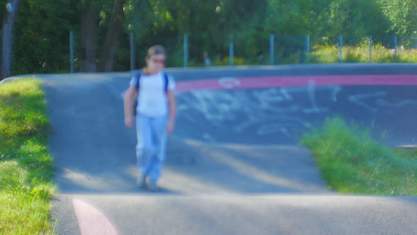 Young girl wearing sunglasses and a backpack walks along the edge of an empty skatepark bowl, enjoying a sunny day outdoors in a vibrant urban setting