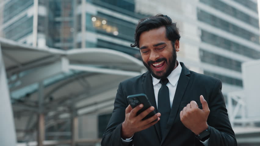 A thrilled businessman stands outside near office buildings, joyfully reading positive news on his smartphone. The excited entrepreneur celebrates success with a smile in a modern business district.