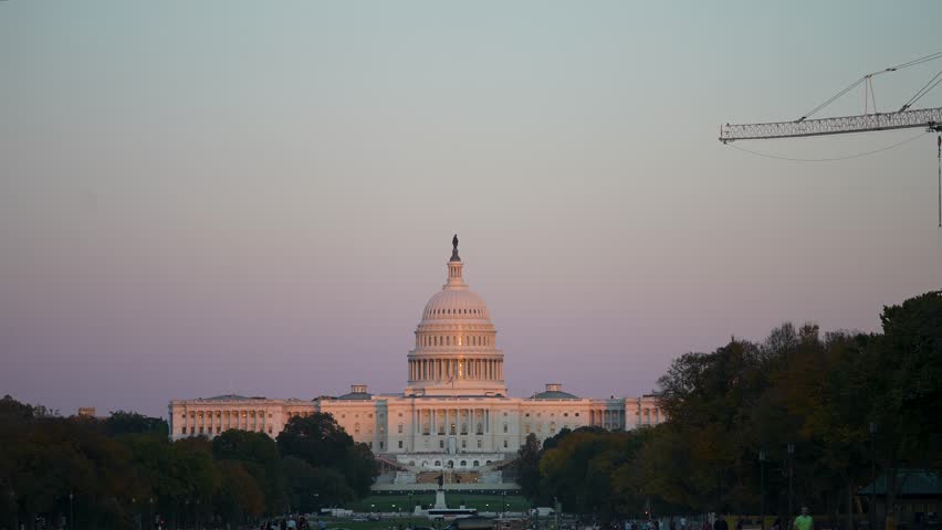 The United States Capitol Building stands tall against a soft pink sunset sky, with a construction crane visible in the upper right corner. Trees line the front of the building, and a few people walk 