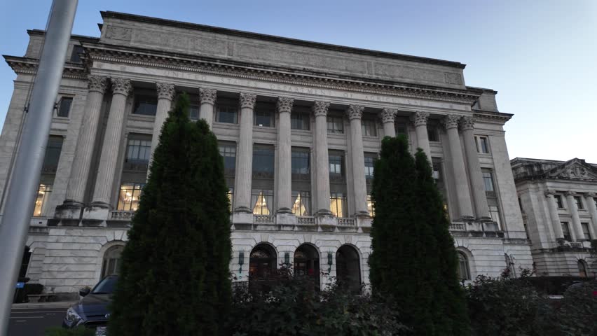 A white marble building with columns, windows, and a sign reading United States Department of Agriculture. There are bushes and trees in front of the building. The sky is blue and there is a flagpole 