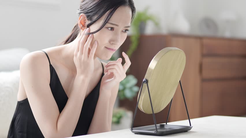 beautiful asian woman using mirror to check skin problem on face in living room