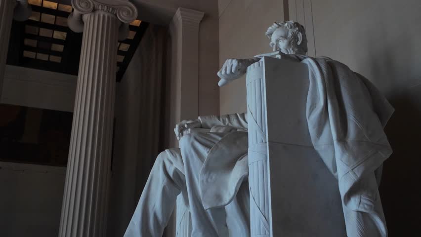 Closeup of Abraham Lincolns marble statue inside the Lincoln Memorial in Washington D.C. He sits with his right arm resting on the chair, his left leg crossed over his right.