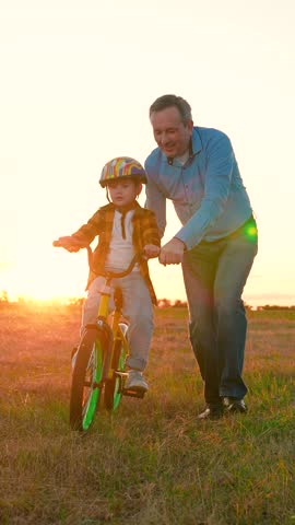 Father teaches his son to ride bicycle on lawn at sunset. Child, dad take walk in park with bicycle in summer. Slow motion. Parent dad little boy in helmet drive bicycle. Sports family weekend dad son