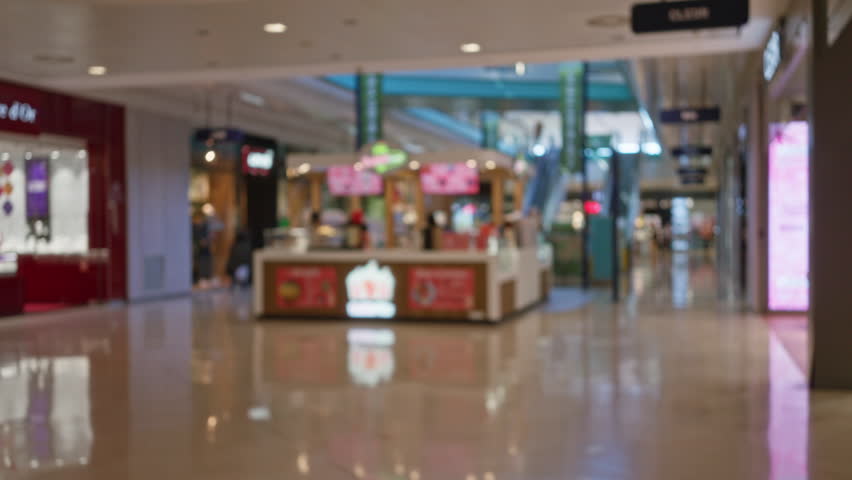 Blurred mall scene with bokeh of indoor shopping center showing kiosks, storefronts, and reflections indicating a bustling commercial environment.