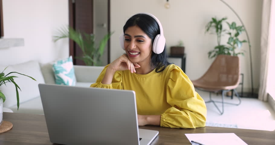 Happy young Indian freelancer woman in headphones talking on video conference chat at laptop, making call from home, speaking at monitor, listening, smiling, enjoying conversation