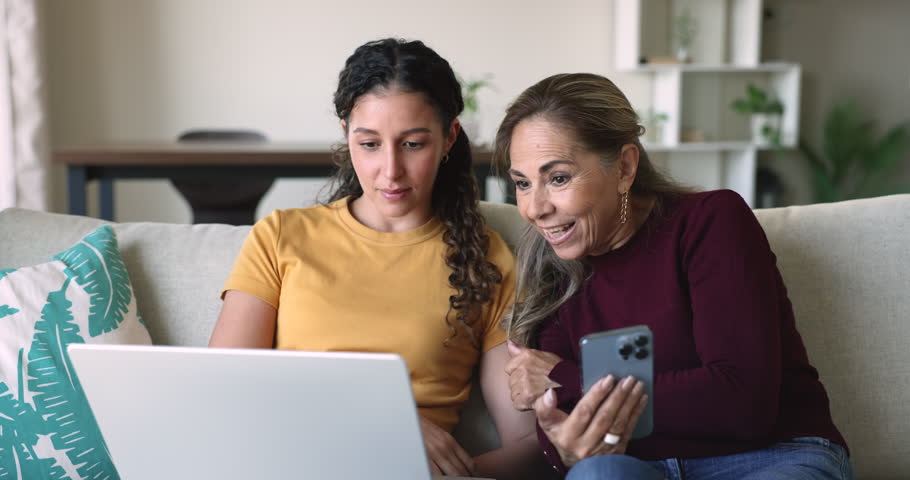 Senior Latin mother and adult daughter girl sharing application, using smartphone and laptop simultaneously, comparing contents on screen and monitor, discussing wireless connection