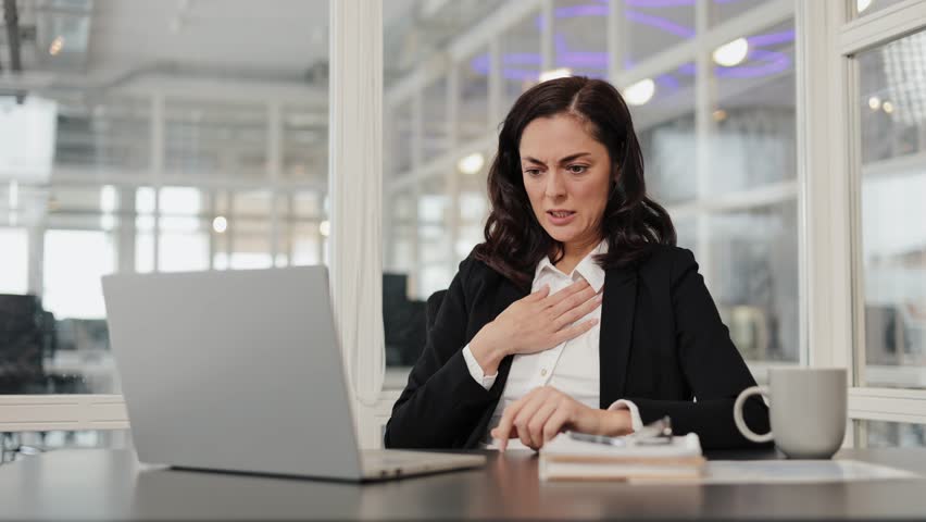 Businesswoman sits at her desk, holding her chest, feeling unwell. Woman has shortness of breath and chest pains, indicating a possible health issue while working.