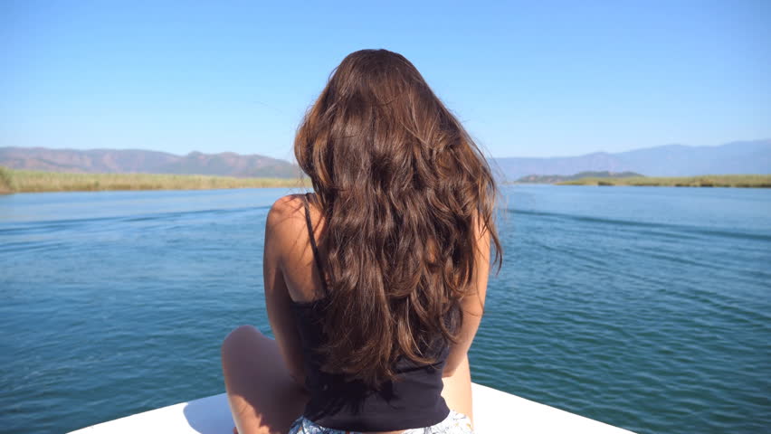 Rear back view of young girl sitting on bow of boat and looking to beautiful nature landscape during trip. Happy woman enjoying summer travel. Vacation or holiday concept. Close up Slow motion
