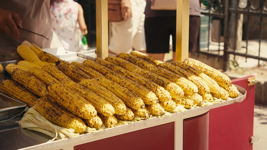 A slow-motion of a street seller roasting corn and many displayed in front of him, with passerby in the background on a sunny day in Istanbul, Turkey