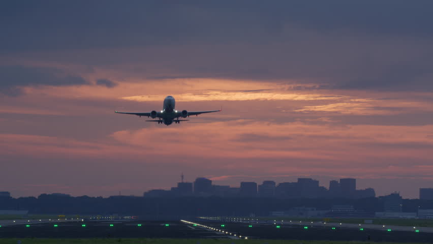 Airplane takes off on a runway with a glowing sunset sky and illuminated runway lights in the background
