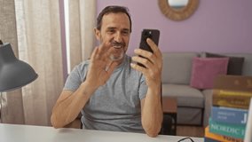 Middle-aged hispanic man in a living room having a video call on his smartphone, smiling and engaging, sitting by a table with books and a lamp in a brightly lit home interior - Powered by Shutterstock - Get 15% off with code: PIKWIZARD15