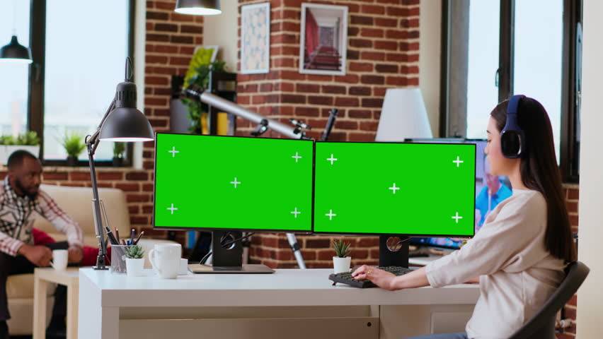 Asian woman solving her freelance tasks with isolated copy space on screen, using headphones while she does research for a project at home office. Remote working and multitasking. Camera B.