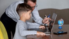 A boy sits at a laptop while his father stands beside him, guiding and explaining something by pointing at the screen. - Powered by Shutterstock - Get 15% off with code: PIKWIZARD15