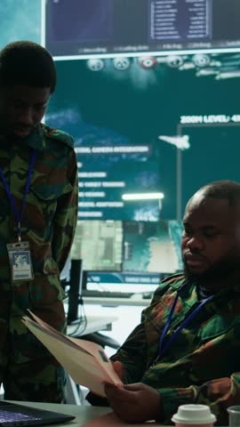 Vertical Video Angry army expert in a command post power base monitors satellite footage, yelling at soldier and throwing papers. Frustrated officer looks at real time data for operational success