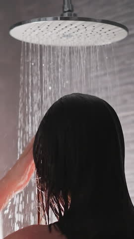 Brunette woman washes hair and body in shower. Lady strokes hair in modern shower with greyish tile. Water from big shower flows down body of lady