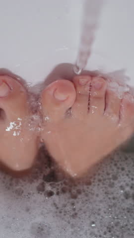 Woman holds feet under running water taking bath at home. Lady massages tired legs with liquid flow in bathroom. Hygiene and relaxation in bath