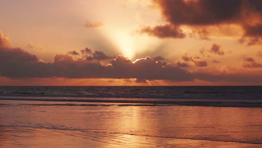 Aerial view flying along the Pacific Ocean coast of Blacks Beach in La Jolla California as the sun sets behind thick clouds creating beautiful colors in the sky as the waves crash on the sandy beach