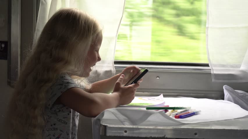 Happy child girl riding in a train carriage near the window and using a smartphone