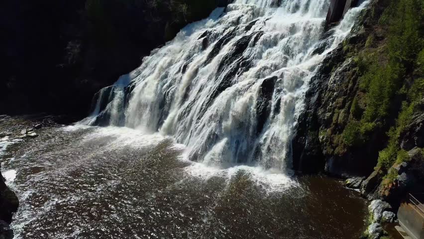 Drone flight to a magnificent waterfall. Climb the falls to its old dam, its metal footbridge and its reservoir that swirls with foam at the top. Riviere-du-Loup, Quebec.