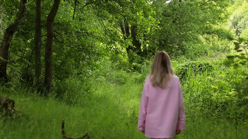 Unrecognisable young Woman in long pink shirt Walking in green Forest and Cat Running next to her. Happy blonde haired Girl Strokes cute Kitten in park outdoors. Slow Living and Relaxation Concept