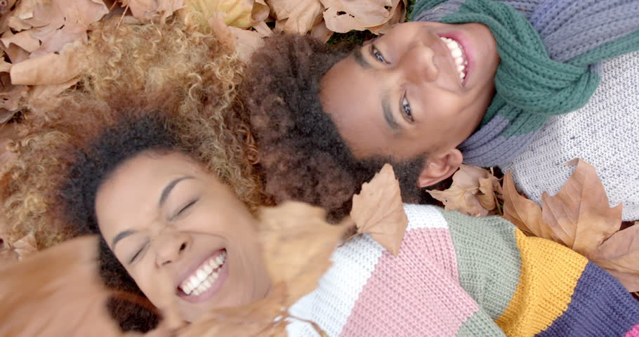 Happy african american mother and son in warm clothes lying on fallen leaves in garden, slow motion. Motherhood, childhood, togetherness, autumn and leisure, unaltered.