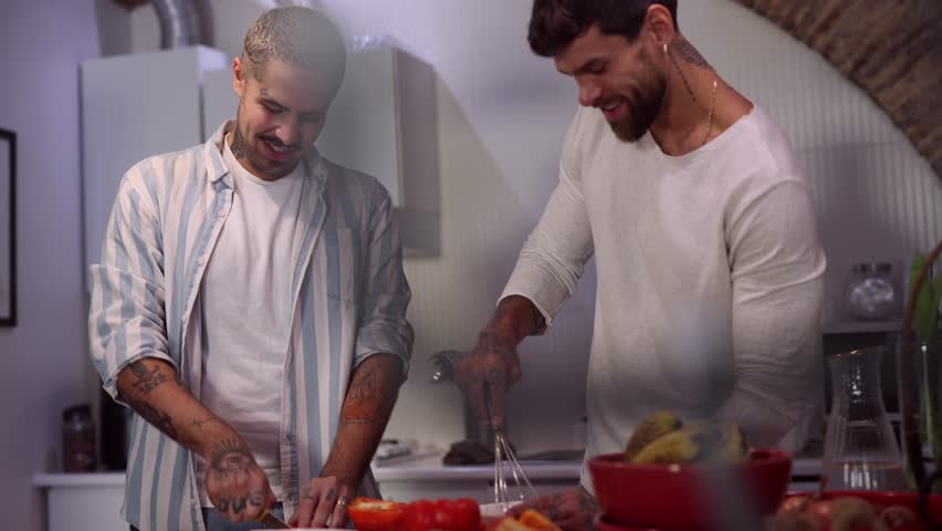 Two men happily cooking together in a modern kitchen, bonding over food and sharing intimate moments. Tattooed arms chopping veggies and cracking eggs
