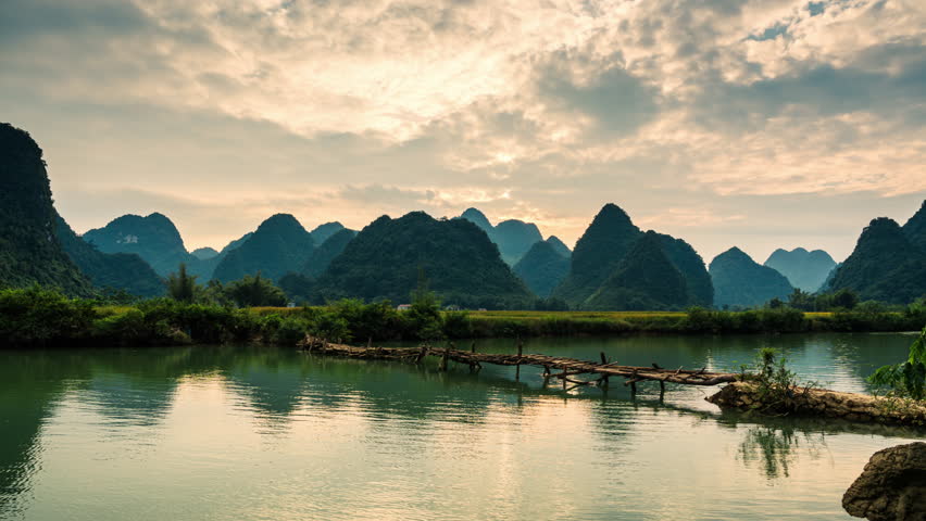 Timelapse of sunset over mountain range with farmer walking on wooden bridge over river in countryside at Phong Nam Valley, Cao Bang, Vietnam