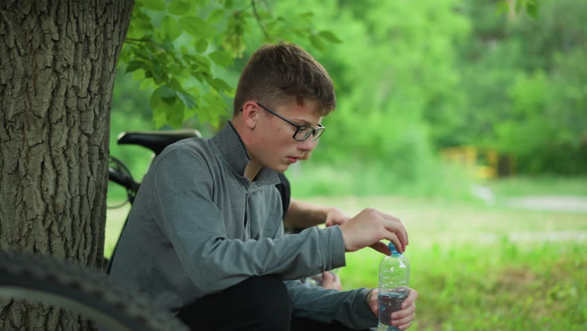 Two young siblings take a break under a tree, one drinks water while the other looks into the distance holding a water bottle, the background features a blur of greenery and natural surroundings