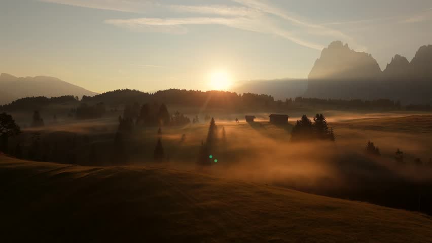 Sunrise over misty meadows in Seiser Alm, Dolomites, captured in slow motion with a drone. Morning light illuminates the valley, highlighting cabins, trees, and mountain peaks in a alpine landscape.