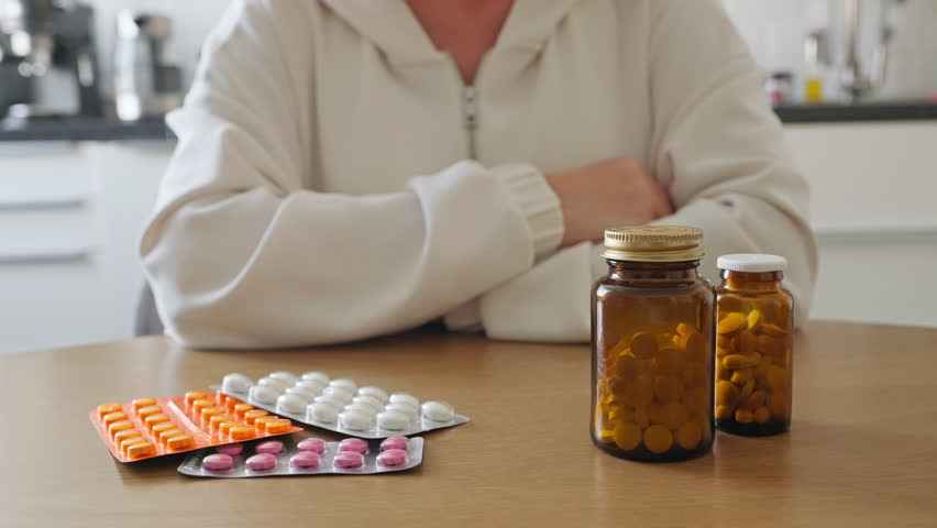 Elderly woman opening brown bottle of pills, surrounded by various tablets blister packs on table in home with kitchen in background. Conveys themes of healthcare, medication management, elderly care.