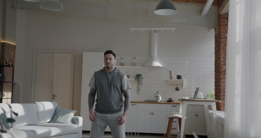 Motivated young man doing sports squatting and jumping enjoying individual training at home. Workout and modern lifestyle concept.