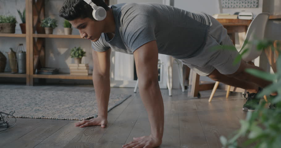 Middle Eastern sportsman doing push-ups and listening to music through wireless headphones indoors at home. Workout and active lifestyle concept.