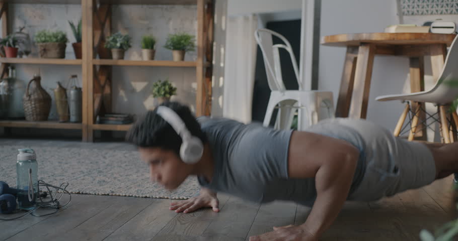 Middle Eastern sportsman doing push-ups and listening to music through wireless headphones indoors at home. Workout and active lifestyle concept.