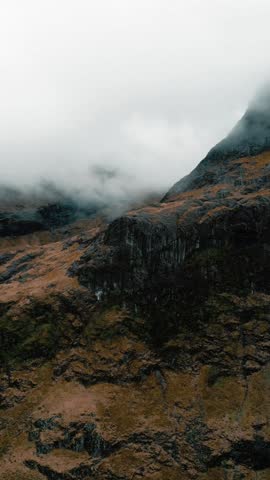 A drone vertical footage of the foggy rugged Scottish Highlands landscape of Glencoe village with mountains, roads and Lagangarbh Hut in Scotland