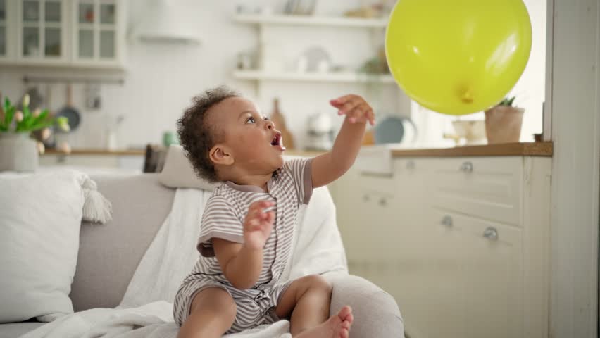 Funny African American toddler playing balloon, portrait of child at cozy home. Funny little black kid sitting on soft sofa in living room and rejoicing, fun and good mood, happy moments of childhood