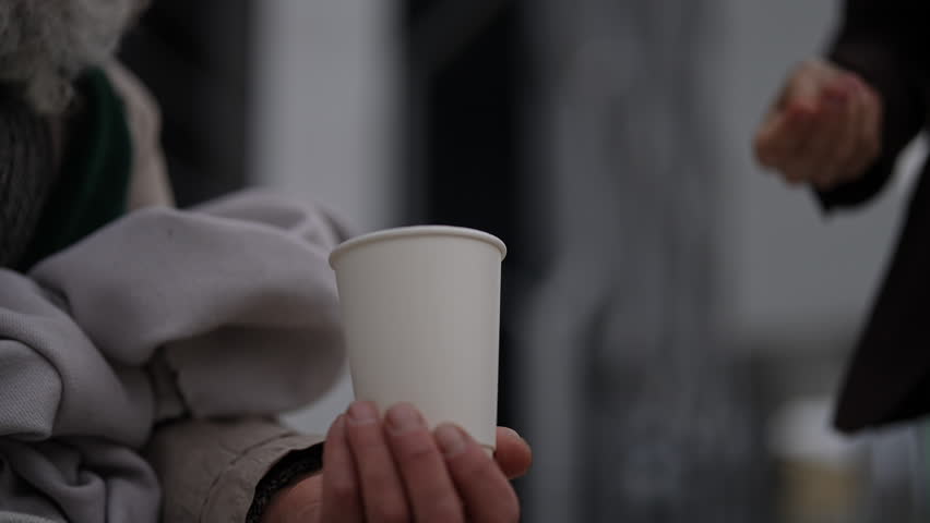 Contrast between wealth and poverty as businessman donating coins to homeless beggar cup in moment of giving and receiving. Closeup hands of business man dropping coins into homeless beggar cup.