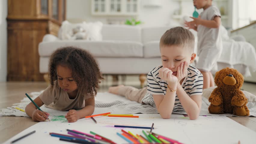 Funny kids playing in playroom, multiracial toddlers drawing by crayons. Childhood and parenting, African American girl and Caucasian boy lying on floor in living room, children at home, cozy interior