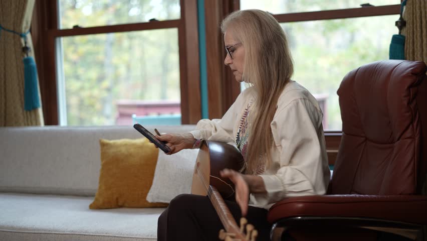 A woman with blonde hair and glasses sits in a brown leather chair, playing a saz instrument. She is wearing a white blouse and black pants. A white couch with yellow pillows is in the background.