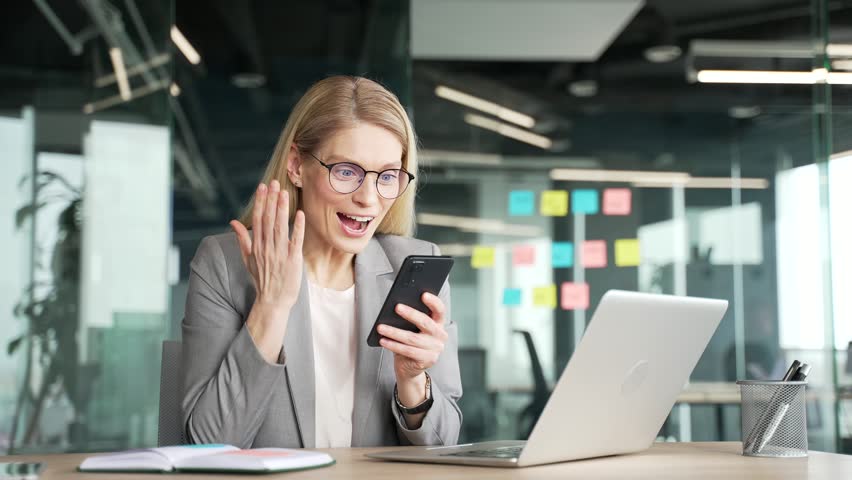 Happy excited businesswoman in jacket reading great news on mobile phone while sitting at desk in business office. Joyful woman celebrates success, smiles, rejoices at positive message on a smartphone