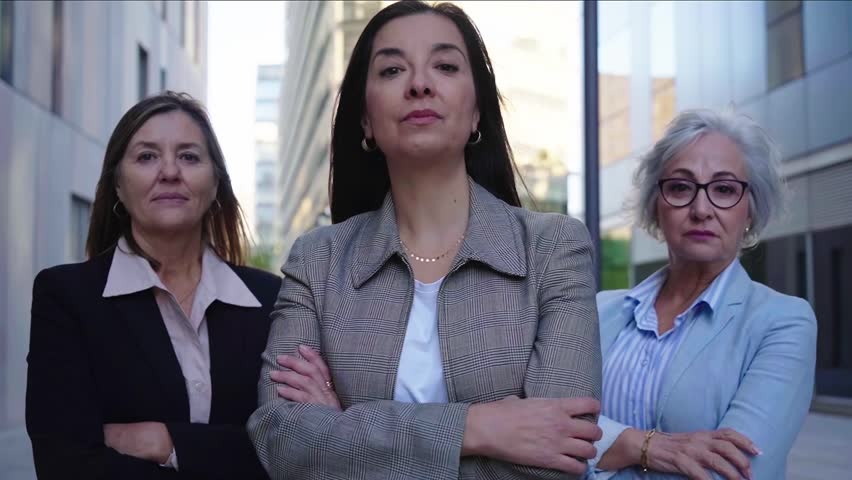 Three mature caucasian empowered businesswomen looking at camera confident. Serious women with cross arms standing outside office district.