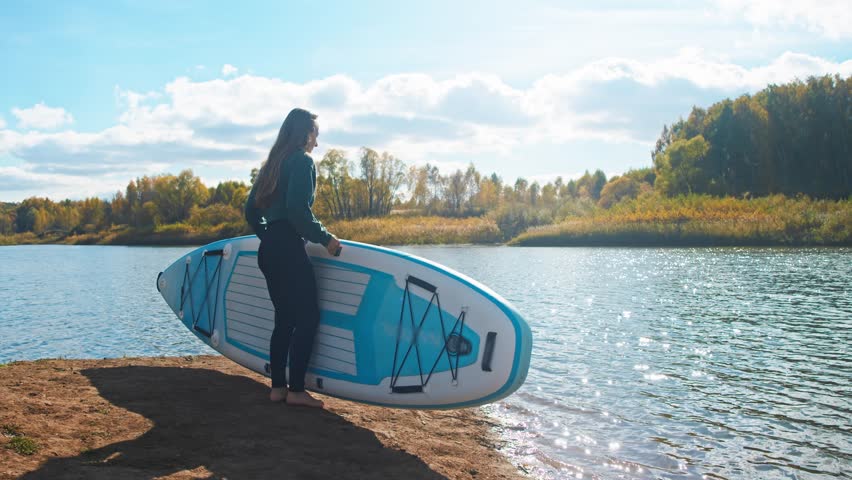 On serene autumnal day, young woman takes to water, launching her stand-up paddleboard in preparation for embarking on her practice of yoga on surface of water.